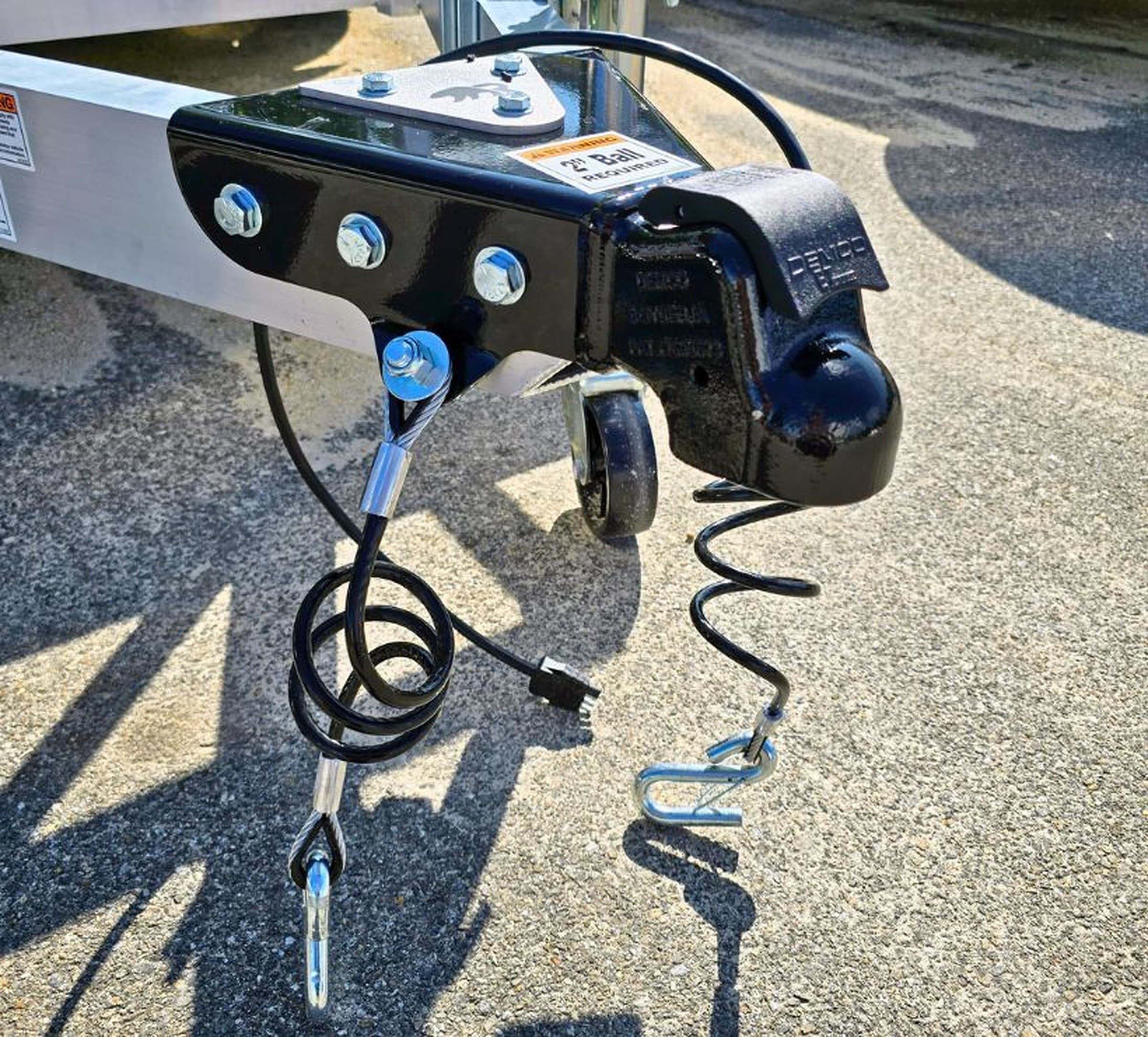 Coupler at the front of a Rugged trailer tongue with crossed safety cables attached to the hitch.